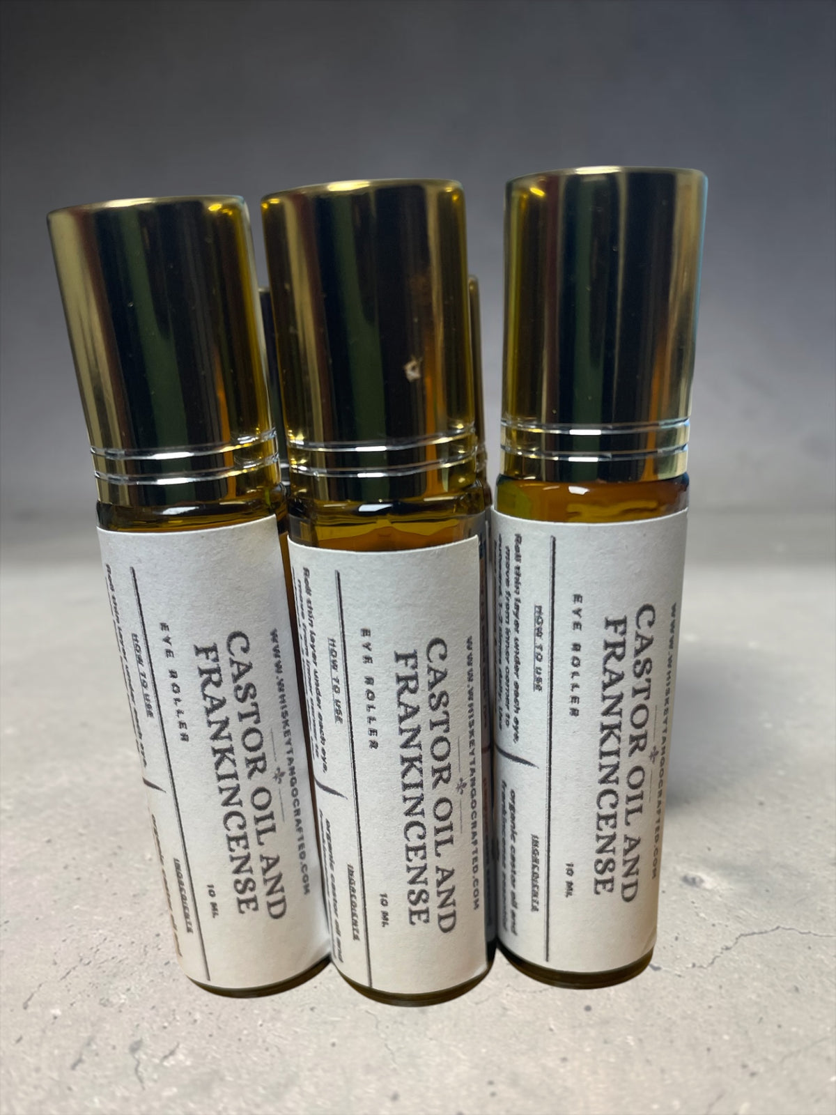 Three amber glass eye roller bottles with white labels and shiny gold caps, labeled 'CASTOR OIL AND FRANKINCENSE EYE DOSSLE'.