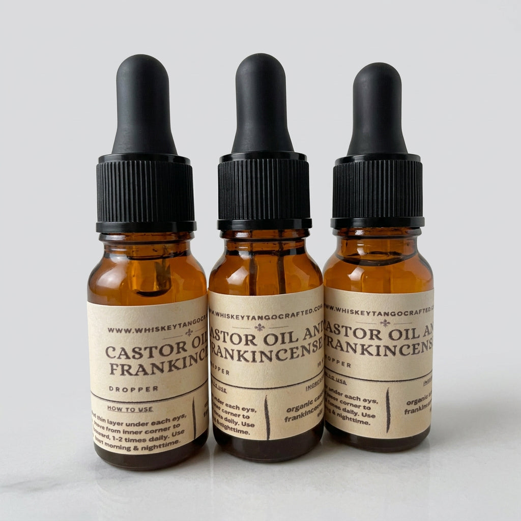 Three amber glass dropper bottles of Whiskey Tangs Castor Oil and Frankincense essential oil, arranged side-by-side on a stone surface with a blurred blue background.