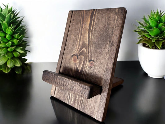 A wooden cookbook stand with a foldable design, displayed in a kitchen setting with two potted plants in the background.
