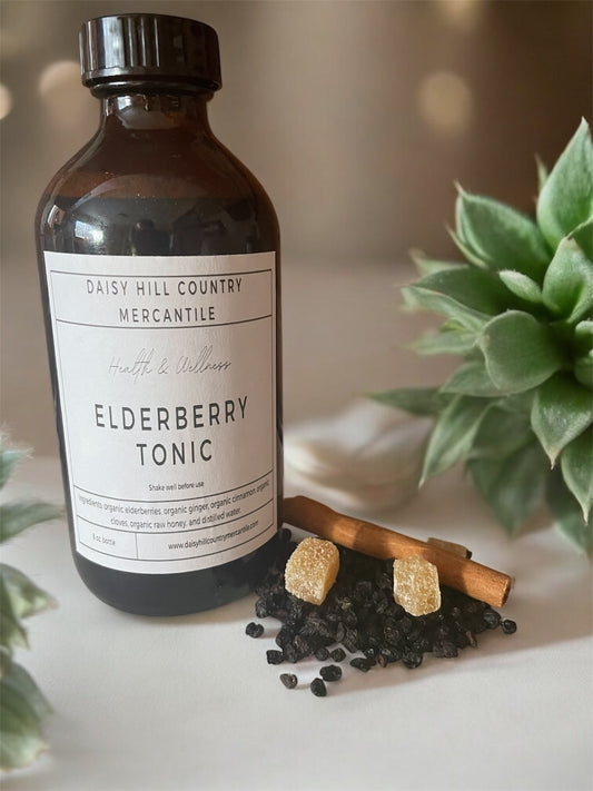 A dark amber glass bottle labeled 'Elderberry Tonic' with a white cap, alongside organic dried elderberries, honey, ginger, cinnamon sticks, and whole cloves, all placed on a white surface with a green plant in the background.