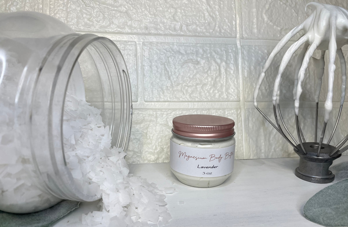 A jar of magnesium body butter with a label that reads 'Magnesium Body Butter'. The jar is placed on a counter next to a whisk, with a white brick wall in the background.