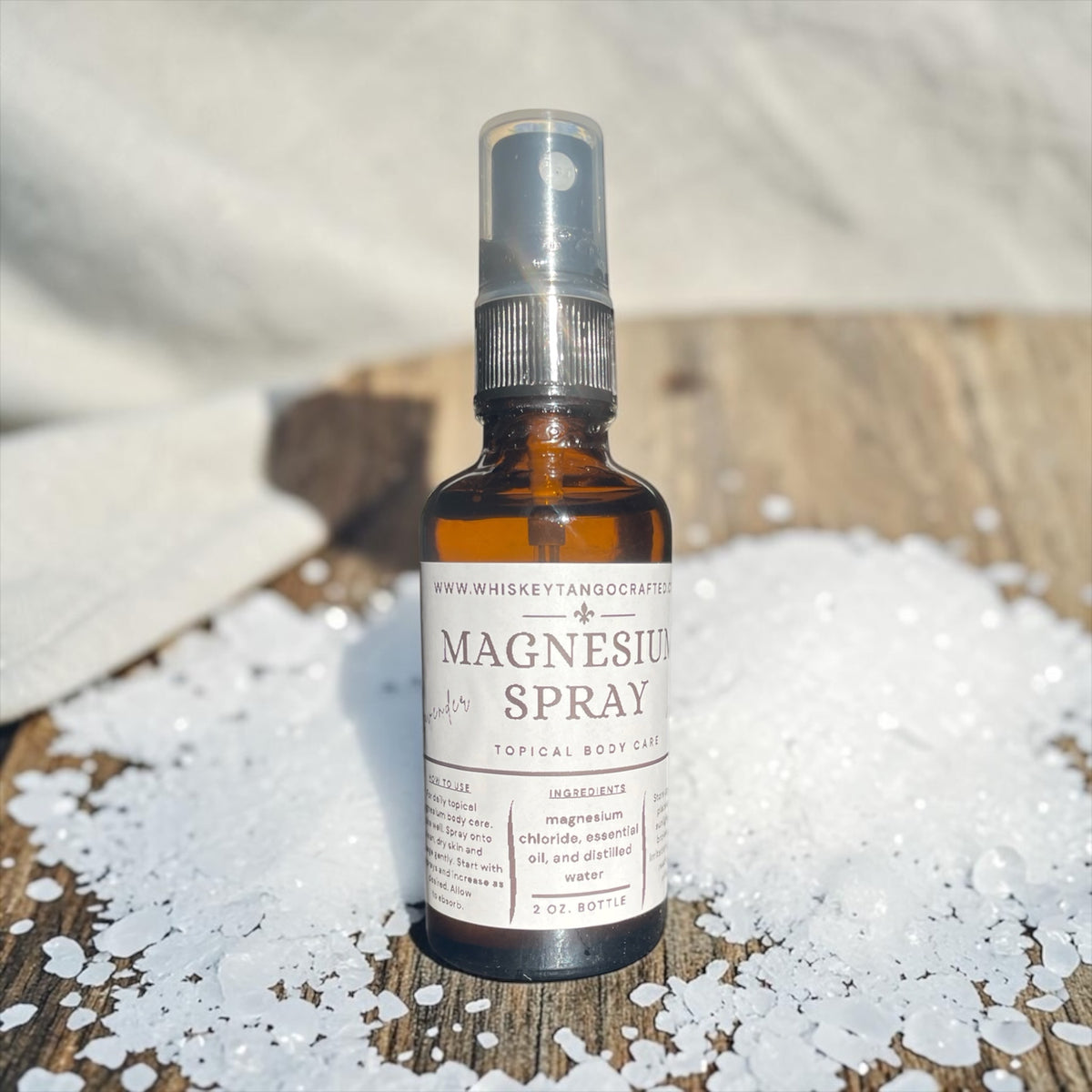 A 7 oz amber glass bottle of Magnuson Spray Topical Body Care, set on a rustic wooden surface surrounded by white sea salt crystals. The background features a soft, draped white fabric.